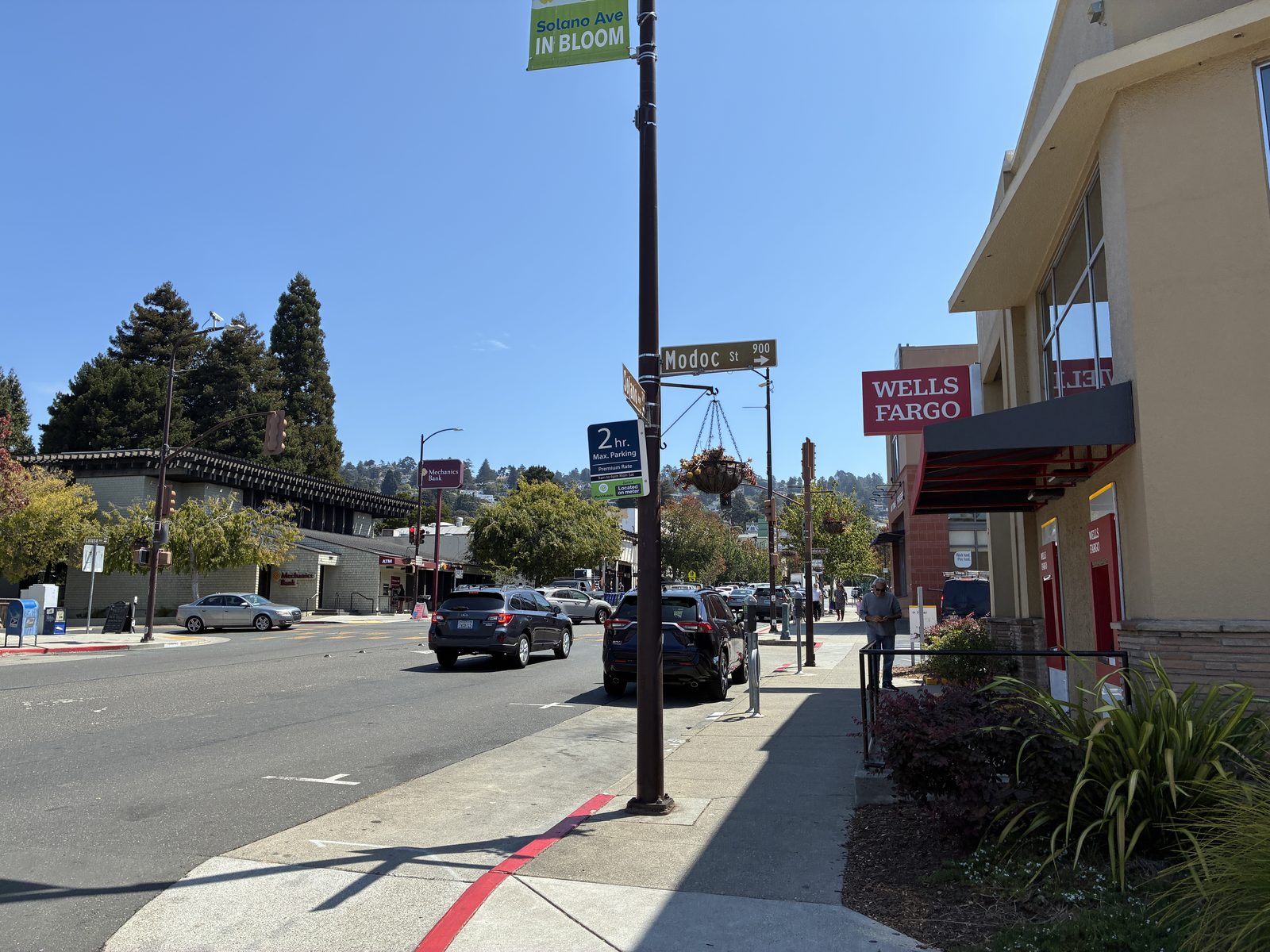 A Solano Avenue sidewalk with a bank branch and a green In Bloom banner overhead
