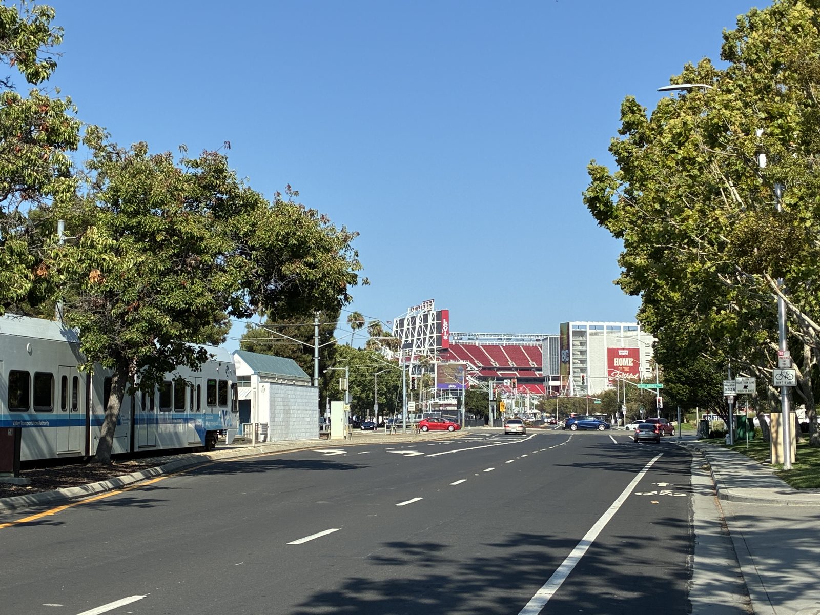 A light rail car moving away down a wide road, the stadium small in the distance
